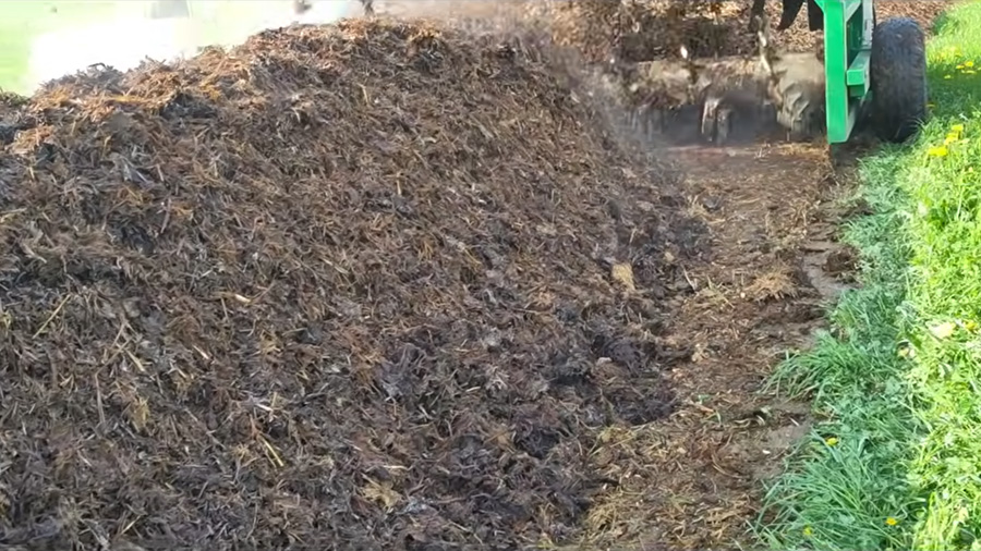 Nahaufnahme der gedrehten Mistmiete mit aufsteigendem Dampf und den Walzen des Gujer Kompostwenders im Hintergrund.