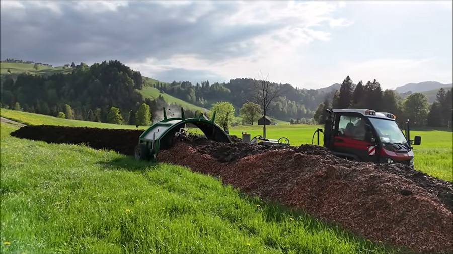 Mistaufbereitung mit Gujer Kompostwender und Aebi Vario VT 450 vor der Mistmiete mit Hackschnitzeln und Mist, umgeben von grünen Wiesen und Hügellandschaft in Bächli-Hemberg.